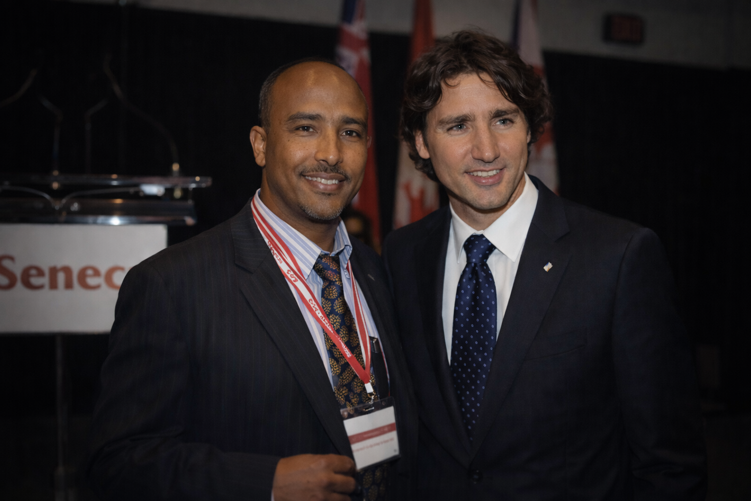 Two men in suits smiling and posing together at an indoor event, one wearing a red lanyard and name badge.