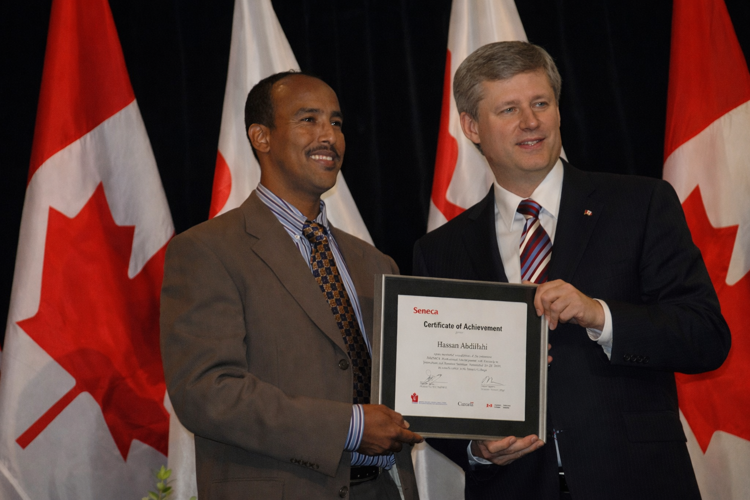 Two men standing in front of Canadian flags, one presenting a Certificate of Achievement to the other.