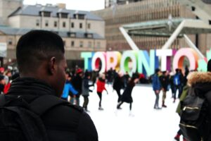 Person in foreground overlooking outdoor ice skating rink with colorful TORONTO sign and people skating in the background.